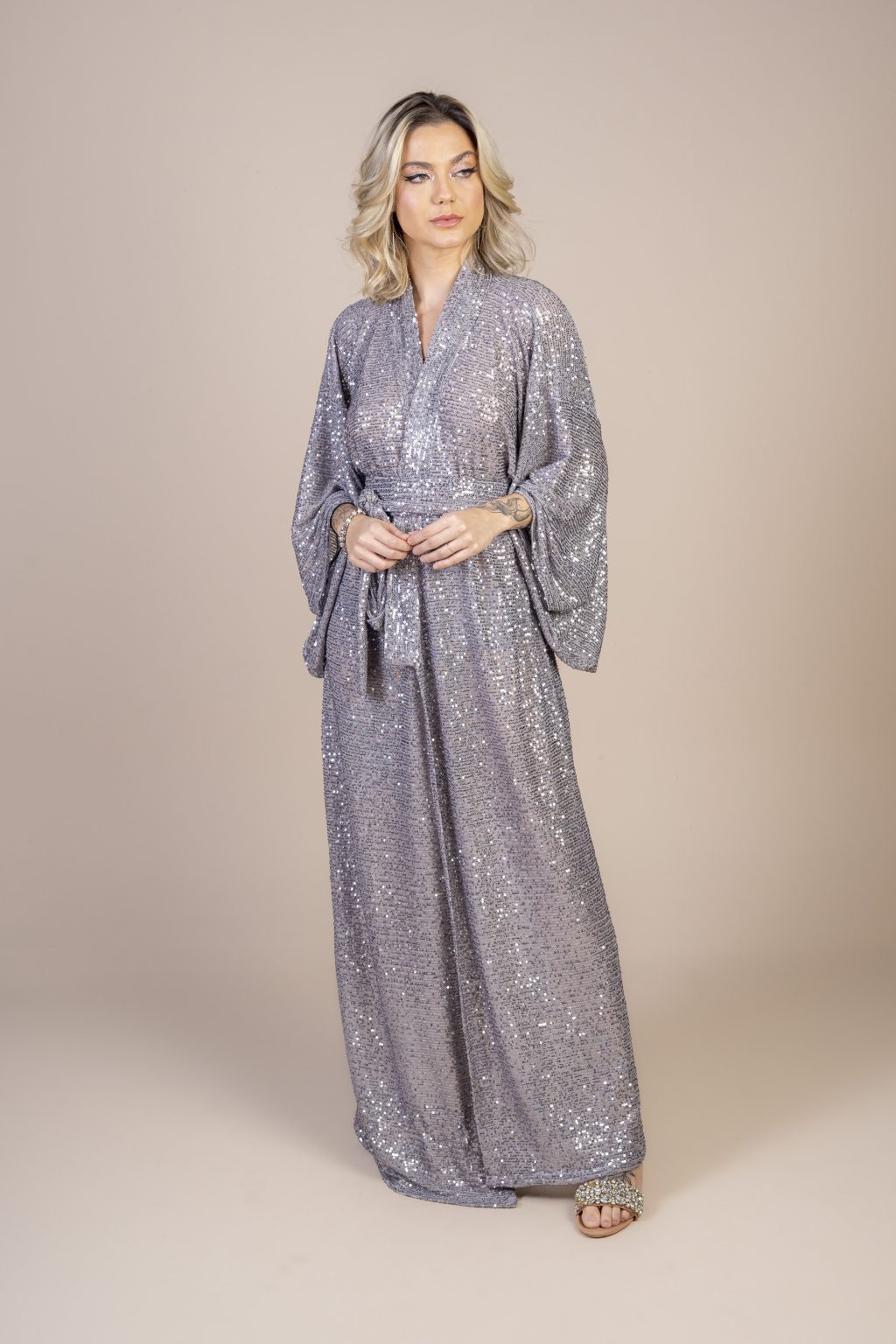 Mulher loira usando um kimono longo manga longa tecido de paetê prata brilhoso faixa de amarração na cintura festa conforto elegância praticidade joker maria sanz kimono quimono